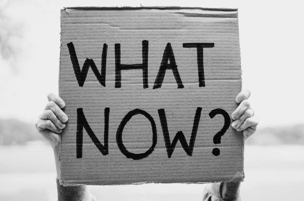 A close-up of hands holding a cardboard sign with the text 'What Now?'